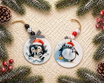 Puede incluir: Dos adornos navideños con diseños de pingüinos. Un pingüino mira hacia adelante, con gorro y bufanda de Papá Noel, el otro mira hacia atrás. Ambos adornos son redondos, con una cuerda para colgar, rodeados de ramas de pino y bayas.
