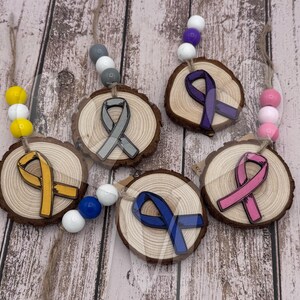 May include: Five wooden ornaments with different colored ribbon designs. The ribbons are gray, purple, yellow, pink, and blue. Each ornament is attached to a string of colorful beads.