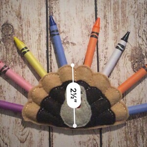 Puede incluir: Una manualidad de pavo de fieltro con un cuerpo marr&oacute;n y plumas negras. El pavo tiene una cabeza gris y un pico rojo. El pavo mide 2 1/2 pulgadas de alto. El pavo est&aacute; rodeado de crayones de varios colores.