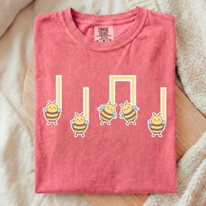 May include: A coral-colored t-shirt featuring a musical note design with cartoon bees. The bees are yellow and brown, with blue wings, and are positioned along the vertical and horizontal lines of the musical note.