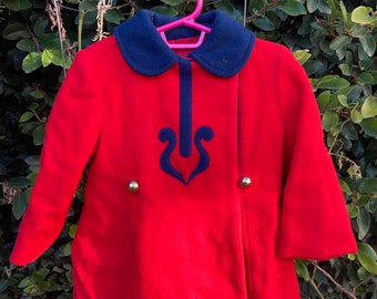 May include: Red wool coat with a blue collar and a blue heart-shaped design on the front. The coat has gold buttons on the front.