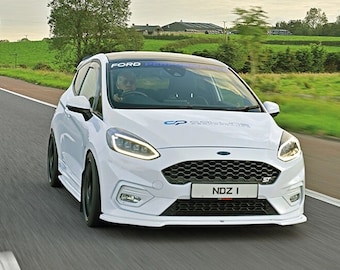 May include: A white Ford Fiesta ST driving on a road with green grass and trees in the background. The car has a black grille, black wheels, and a black spoiler. The license plate reads "NDZ 1".