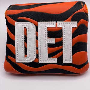 May include: Golf club head cover with a tiger stripe pattern in orange and black. The cover features the letters "DET" in white, stitched onto the front. The cover is designed to protect golf clubs.