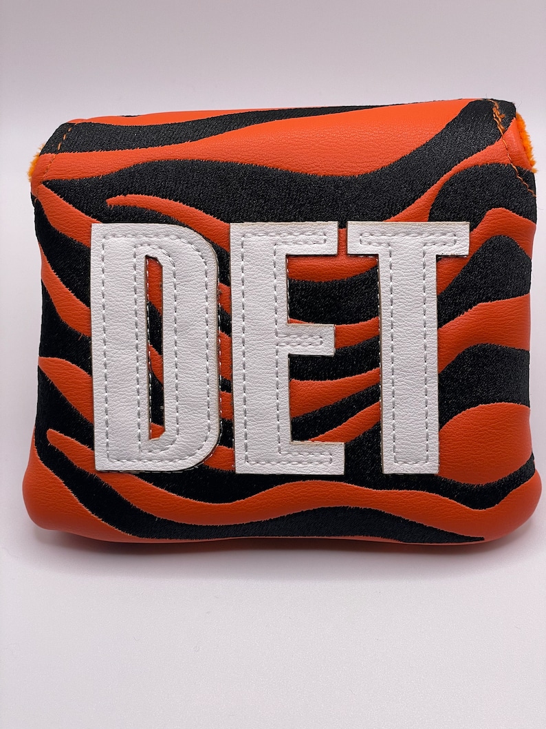 May include: Golf club head cover with a tiger stripe pattern in orange and black. The cover features the letters "DET" in white, stitched onto the front. The cover is designed to protect golf clubs.