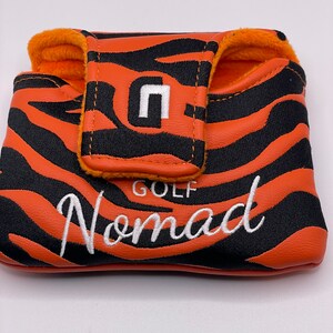 May include: Orange and black golf putter cover with a tiger stripe pattern. The cover has the word "Nomad" embroidered in white script. The top flap features a white "n" logo and the word "GOLF". The interior is lined with orange plush material.
