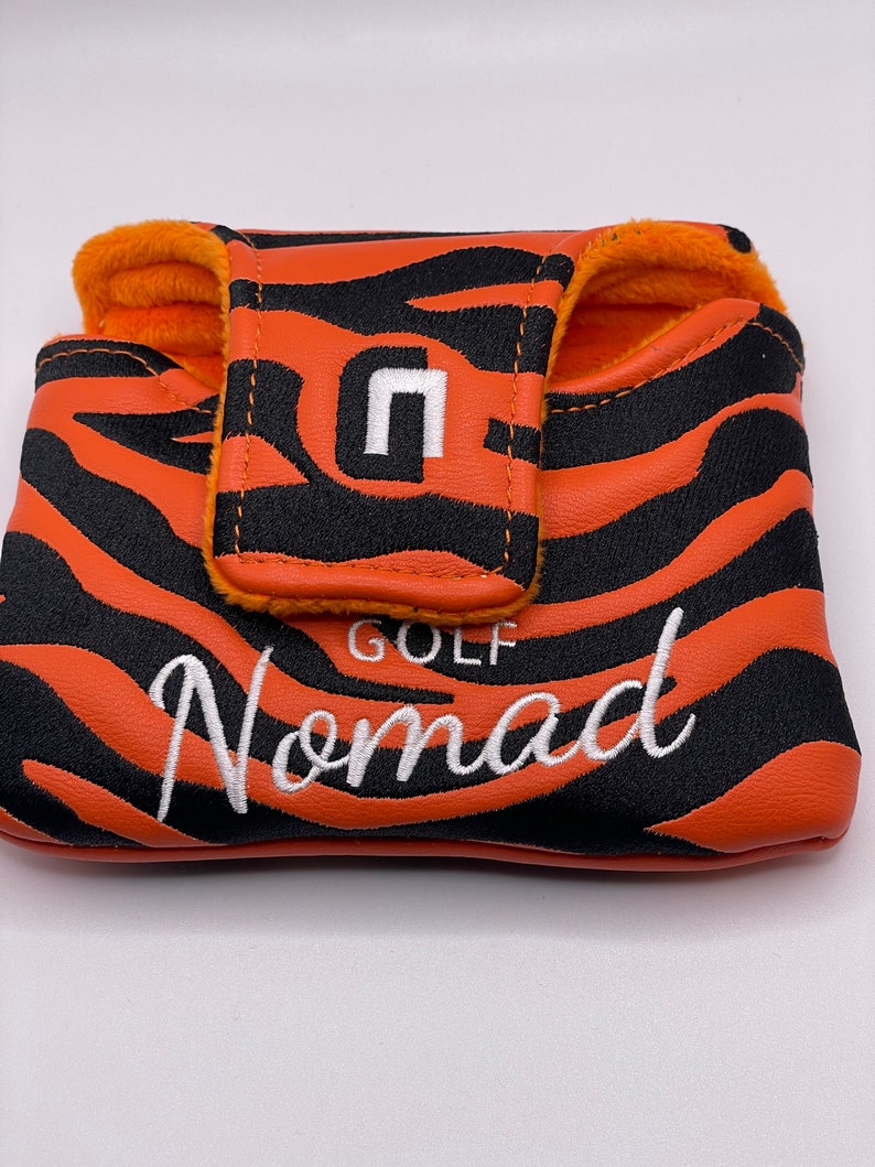 May include: Orange and black golf putter cover with a tiger stripe pattern. The cover has the word "Nomad" embroidered in white script. The top flap features a white "n" logo and the word "GOLF". The interior is lined with orange plush material.