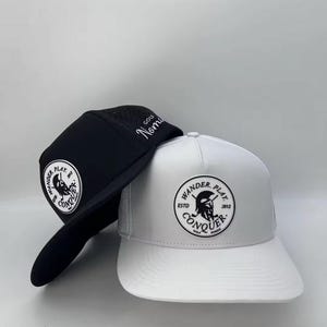 May include: Two baseball caps, one black and one white. The white cap has a black logo with the text "Wander Play Conquer" and the year "2012". The black cap has the same logo in white and the text "Golf Nomads".