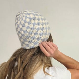 May include: A knit beanie featuring a checkered pattern in soft blue and cream colors. The hat is constructed with a textured yarn and has a rounded crown shape that fits snugly. The design consists of alternating light blue and cream squares.