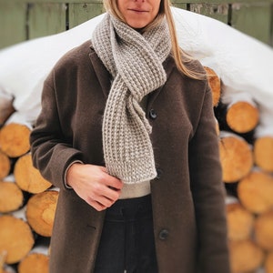May include: A woman wearing a brown coat and a light brown knitted scarf. The scarf is wrapped around her neck and tied in a loose knot. She is standing in front of a pile of logs.