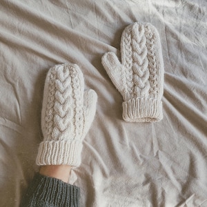 Op de afbeelding: Een paar witte gebreide wanten met een kabelbreipatroon. De wanten liggen op een beige stoffen oppervlak.