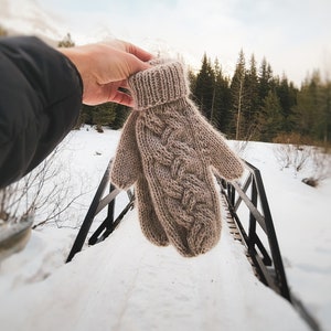 May include: A close-up of a brown knitted mitten with a cable knit pattern. The mitten is held up in the air against a snowy background with a wooden bridge in the foreground.