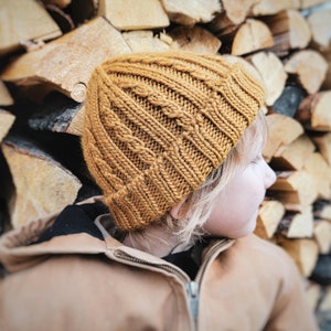 May include: A child wearing a brown knitted hat with a cable knit pattern, sitting against a stack of firewood.