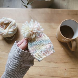 May include: A small, hand-knitted hat in pastel colors of pink, blue, and yellow, topped with a fluffy white pom-pom. The hat is held by a person wearing a gray sleeve. A ball of yarn and a mug of coffee are in the background.