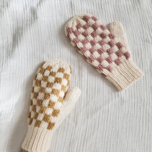 Peut inclure: Deux moufles tricotées avec un motif en damier. L'une présente un motif à carreaux marron et crème, l'autre un motif rose et crème. Les deux moufles ont des poignets crème et sont présentées sur une surface blanche texturée.