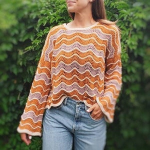 Peut inclure: Un pull en crochet avec un motif ondulé dans les tons orange, rose et blanc. Le pull a une coupe ample et des manches longues.
