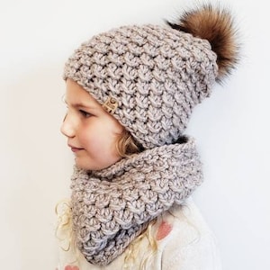 May include: A child wearing a light brown knitted hat with a fluffy brown pom pom and a matching light brown knitted scarf. The hat has a small brown label with the word "Made" on it.