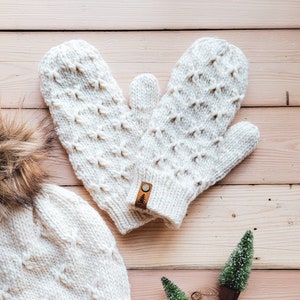 May include: A pair of white knitted mittens with a textured pattern. The mittens have a brown leather tag with a small tree design. The mittens are lying on a light wood surface.