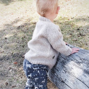 May include: A toddler wearing a beige knitted sweater and blue pants with a white deer print stands next to a large log.