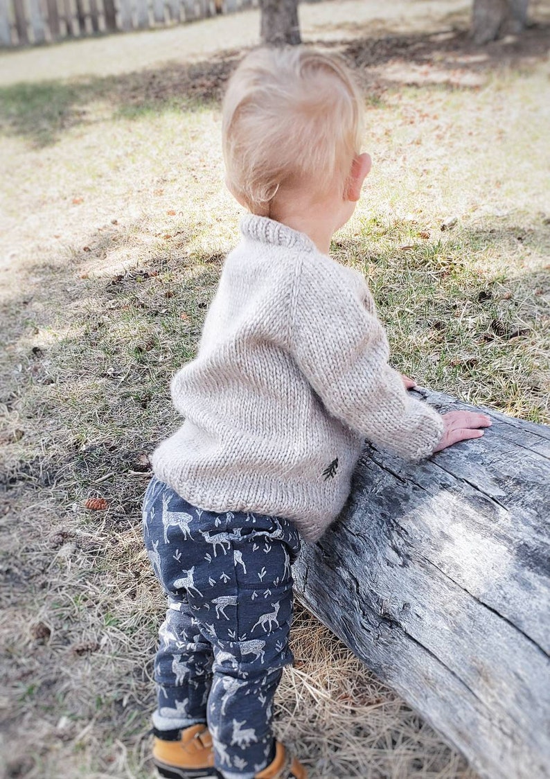May include: A toddler wearing a beige knitted sweater and blue pants with a white deer print stands next to a large log.