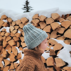 May include: A child wearing a light blue knitted beanie and a brown fleece jacket stands in front of a stack of firewood covered in snow.
