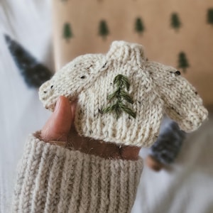May include: A miniature cream-colored knitted sweater with a green embroidered Christmas tree design. The sweater is held in a hand wearing a matching cream-colored knitted sleeve. The background includes a gift-wrapped package with green tree patterns.
