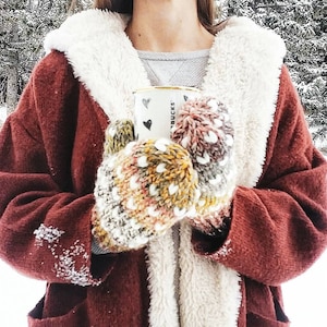 May include: A person wearing a burgundy coat with a white fluffy collar and holding a white mug with black hearts and the word "BUCKS". The person is wearing colorful knitted mittens with white heart patterns.