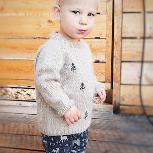 May include: A young child wearing a beige knitted sweater with embroidered pine trees and blue pants with a white deer pattern. The child is standing in front of a wooden wall.