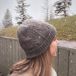 May include: A close-up of a person wearing a gray knitted beanie hat. The hat has a textured pattern and is pulled down over the person's forehead. The person's hair is long and brown, and they are looking off to the side.