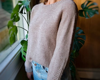 May include: A light brown knitted sweater with long sleeves and a crew neckline. The sweater is worn over a pair of blue jeans.