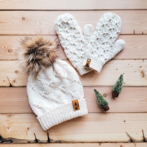 May include: A white knitted hat with a fluffy pom pom and matching mittens. The hat and mittens have a diamond pattern and a small brown leather patch with a tree design.