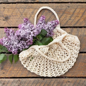 May include: A cream-colored, open-weave knitted tote bag with a handle, filled with vibrant purple lilac blossoms and green leaves. The bag rests on a rustic wooden surface, showcasing a natural, handcrafted aesthetic. The bag's design is open and airy.