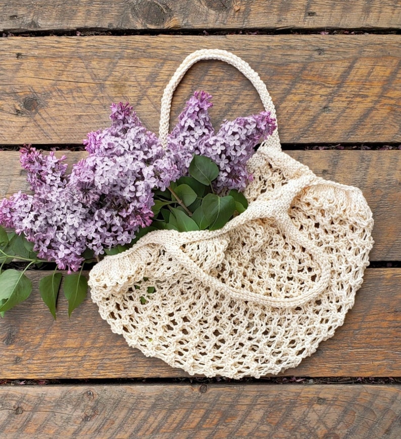 May include: A cream-colored, open-weave knitted tote bag with a handle, filled with vibrant purple lilac blossoms and green leaves. The bag rests on a rustic wooden surface, showcasing a natural, handcrafted aesthetic. The bag's design is open and airy.