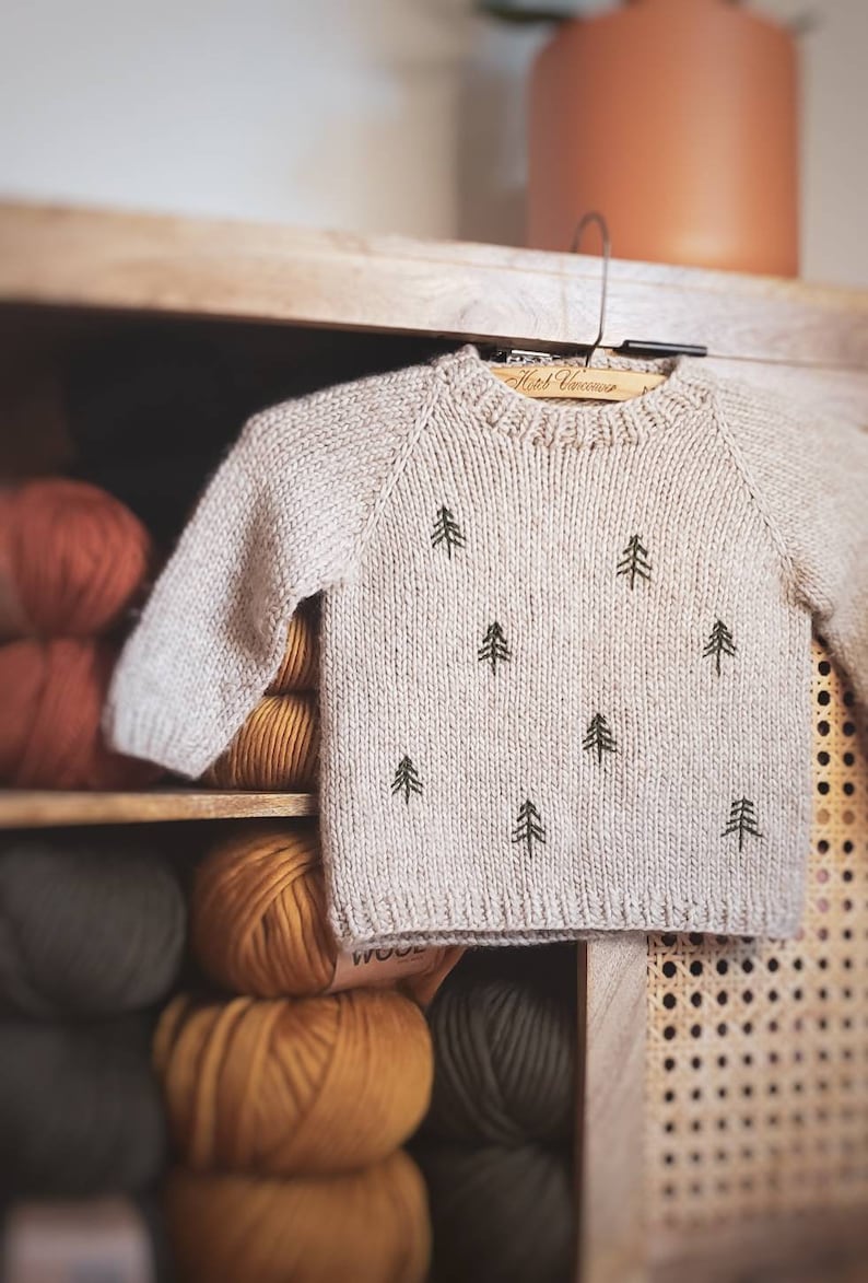 May include: A beige knitted sweater with a crew neck and a pattern of small green trees. The sweater is hanging on a wooden hanger and is surrounded by balls of yarn in various colors.