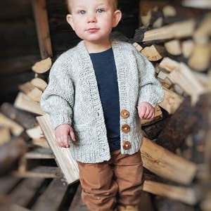 May include: A young child wearing a light grey knitted cardigan with brown wooden buttons. The cardigan has a pocket on the front. The child is wearing brown trousers.