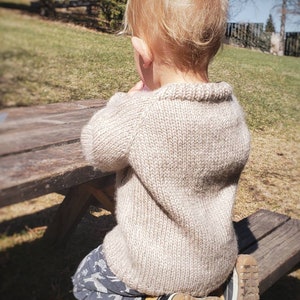 May include: A young child wearing a beige knitted sweater sits on a wooden bench. The child is wearing blue and white patterned pants and brown shoes.