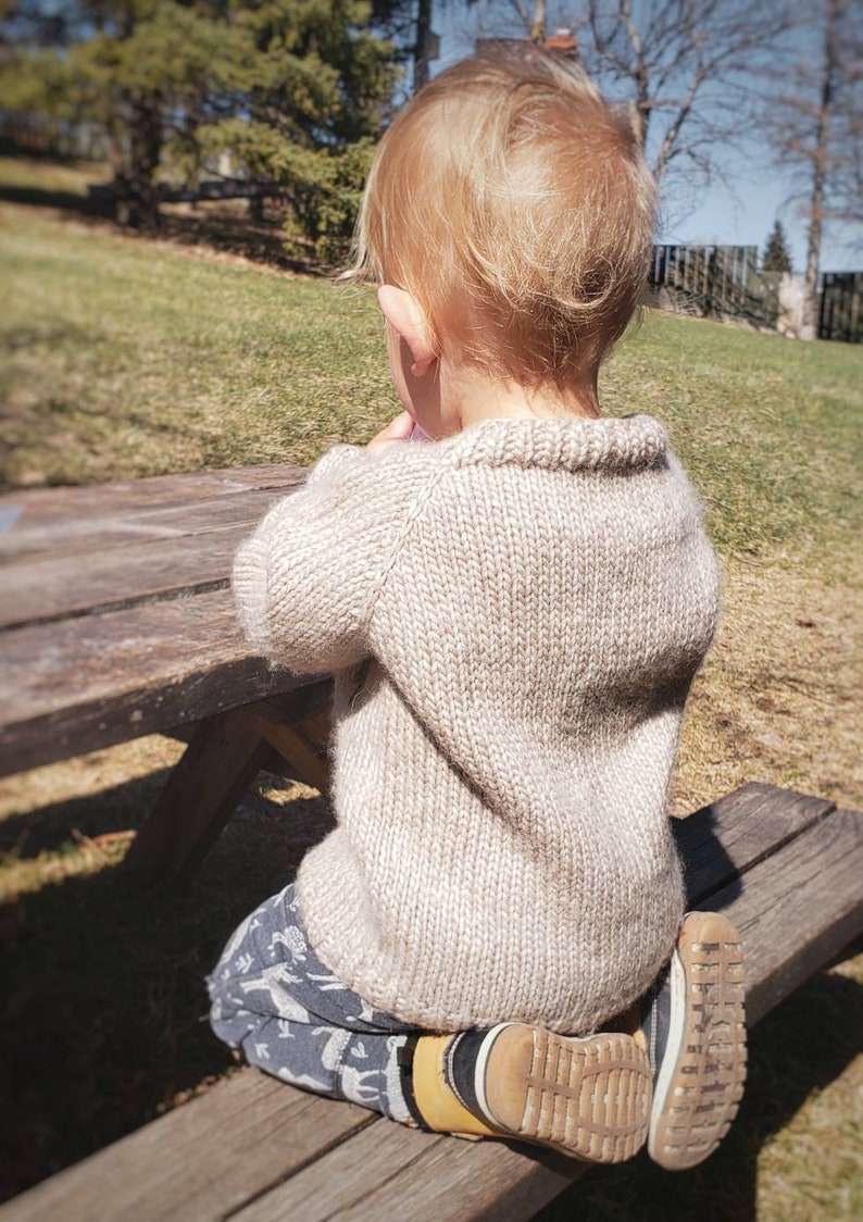 May include: A young child wearing a beige knitted sweater sits on a wooden bench. The child is wearing blue and white patterned pants and brown shoes.