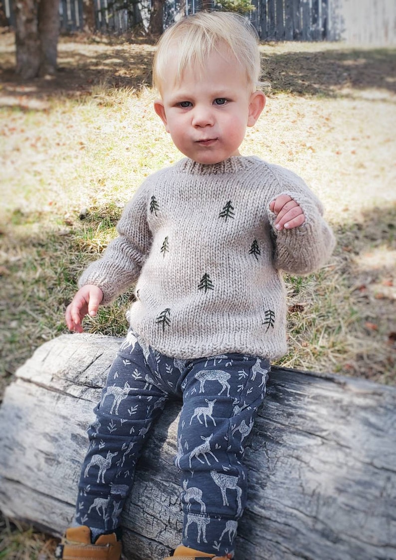 May include: A young child wearing a beige knitted sweater with a pattern of small pine trees and blue pants with a white deer pattern. The child is sitting on a log.