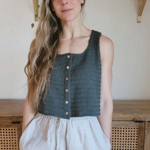 Puede incluir: Un top de punto verde azulado oscuro con botones de madera. El top tiene un escote cuadrado y se combina con pantalones de lino gris claro. El conjunto es informal y cómodo, adecuado para el uso diario.