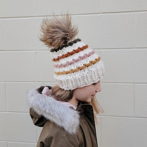 May include: A white knitted beanie with a multi-coloured striped pattern and a brown faux fur pom pom.