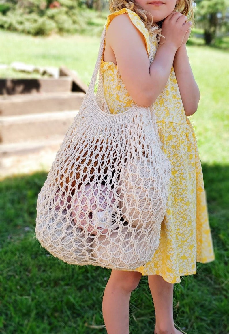 May include: A cream-colored, open-weave tote bag filled with yarn. The bag is carried by a person wearing a yellow floral dress. The bag's design features a large, open netting, revealing the colorful yarn inside. The scene is set outdoors with green grass and a staircase in the background.