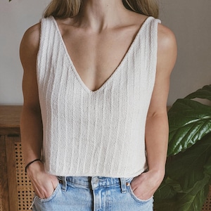 Könnte beinhalten: Ein weißes Strick-Tanktop mit tiefem V-Ausschnitt. Das Tanktop ist aus einem weichen, strukturierten Stoff gefertigt und hat eine lockere Passform. Das Model trägt das Tanktop zu einer blauen Jeans.