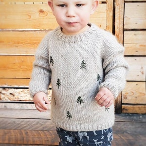 May include: A young child wearing a light gray knitted sweater with green embroidered pine trees and blue pants with a white deer print. The child is standing in front of a wooden wall.