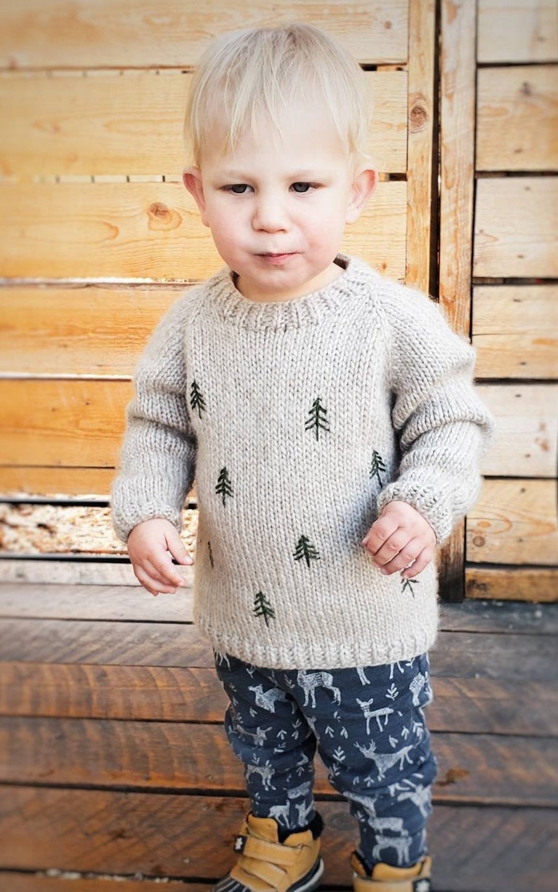 May include: A young child wearing a light gray knitted sweater with green embroidered pine trees and blue pants with a white deer print. The child is standing in front of a wooden wall.