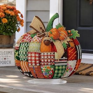 May include: A decorative pumpkin ornament covered in multicolored rhinestones arranged in a patchwork pattern. The pumpkin features a green stem, a gold bow, and small orange accents, all mounted on a clear circular base for stable display.
