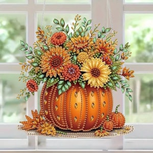 May include: A hanging suncatcher featuring a large orange pumpkin base adorned with embedded crystals, topped with a vibrant arrangement of sunflowers, orange blossoms, and green leaves. Two smaller pumpkins sit at the base of the display.