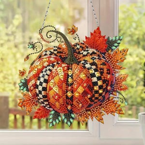 May include: A hanging pumpkin decoration covered in faceted orange, yellow, and black beads arranged in a checkered and diamond pattern. The pumpkin is surrounded by beaded autumn leaves in shades of red, orange, and green, suspended by a thin chain.