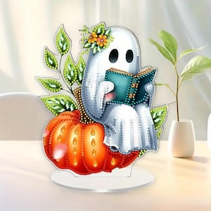 May include: A Halloween-themed decorative piece featuring a white ghost reading a teal book while seated on an orange pumpkin. The ghost is adorned with a floral crown and sits in front of green leaves. The piece is on a clear base.