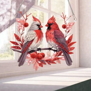 May include: A decorative hanging featuring two cardinal birds perched on a branch. The birds are primarily red and gray, with intricate detailing. The branch is surrounded by red and pink leaves and berries, with a window in the background.