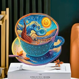 Könnte beinhalten: Ein dekoratives Tassen- und Untertassen-Set mit einer künstlerischen Darstellung von Van Goghs "Sternennacht". Die blaue Tasse hat Sterne, und die Untertasse enthält einen Löffel und Blumendekorationen. Das Set wird auf einem weißen Buch präsentiert.