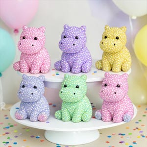 May include: Six small hippopotamus figurines covered in sparkling rhinestones, arranged in two rows. The top row features pink, purple, and yellow hippos, while the bottom row displays light blue, light green, and light pink hippos with black eyes.